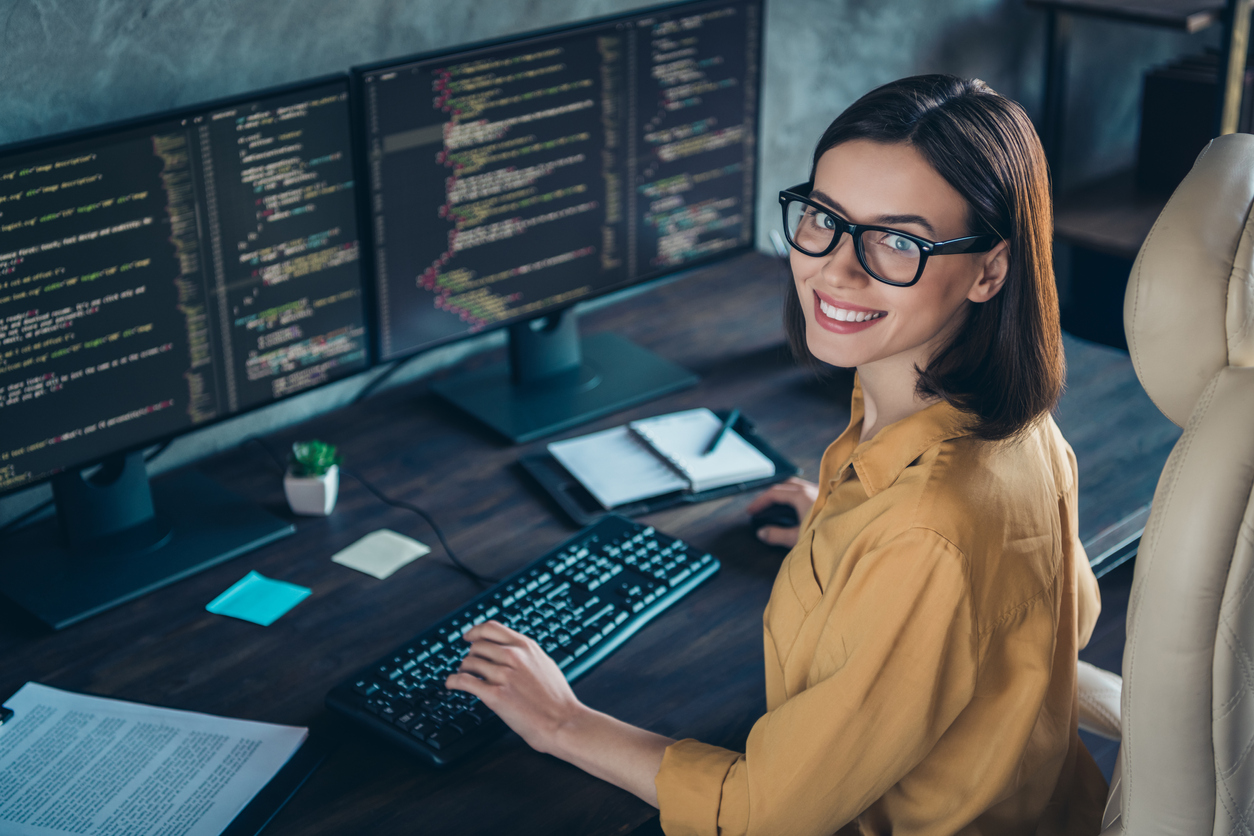 Smiling software developer in glasses coding at dual monitors in a modern workspace.